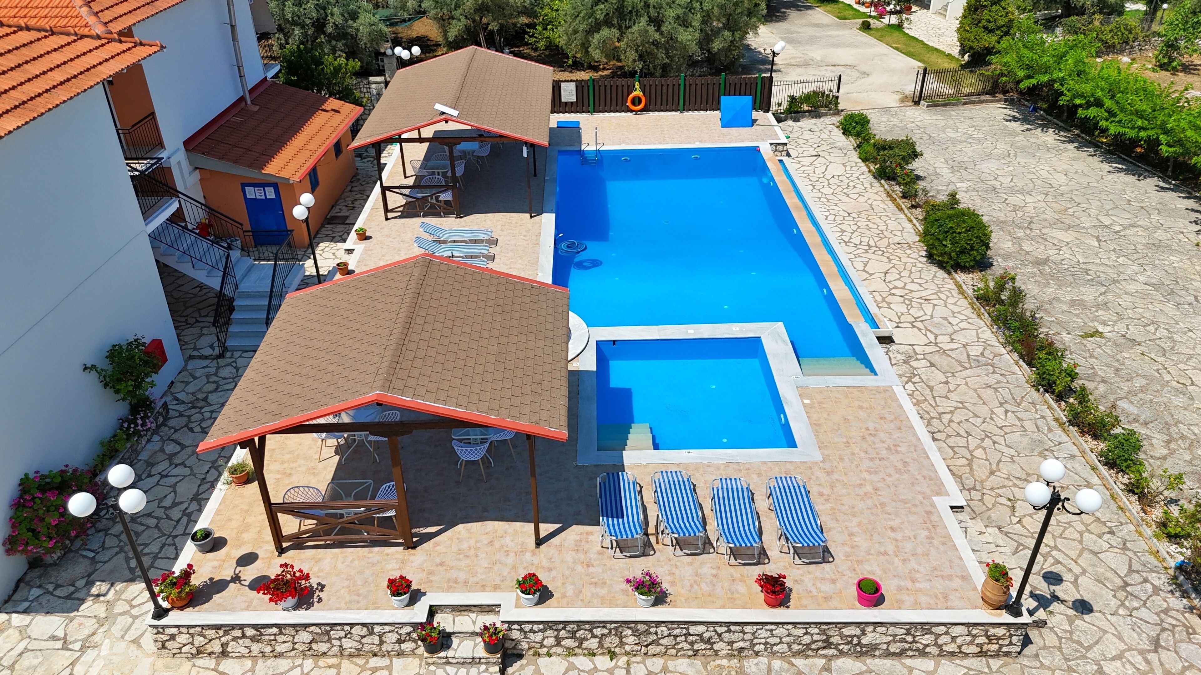 A swimming pool with multiple sections, surrounded by stone-tiled decking. There are lounge chairs and a covered seating area with a red roof.