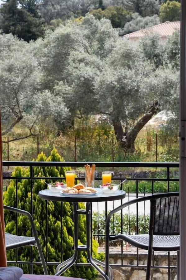 Un balcon avec une table dressée pour le petit-déjeuner, comprenant des croissants et du jus d’orange, donnant sur un jardin avec des arbres et un mur de pierre.