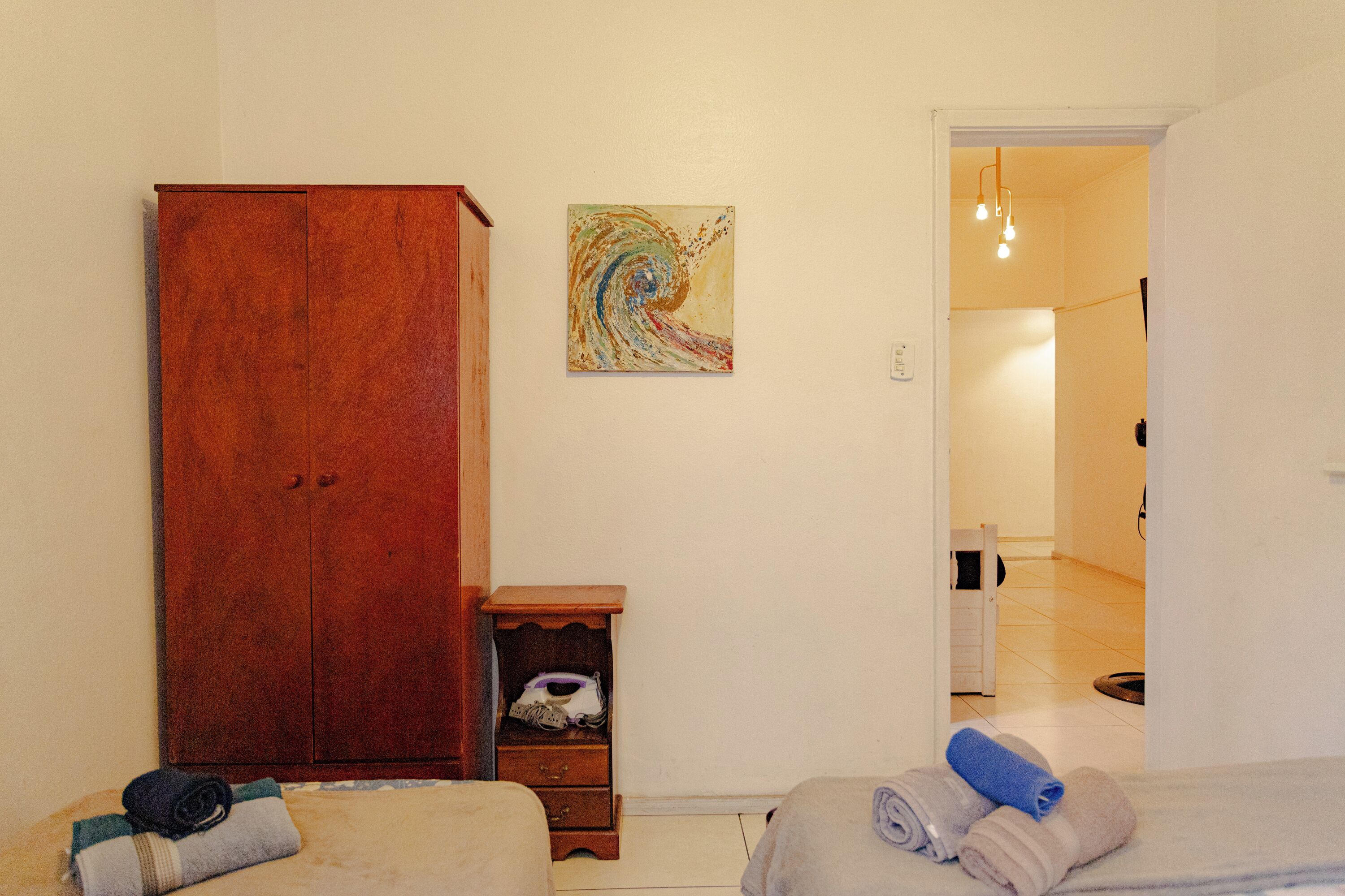 Un dormitorio con un armario de madera, una mesita de noche pequeña, un cuadro en la pared y un baño visible a través de una puerta abierta.