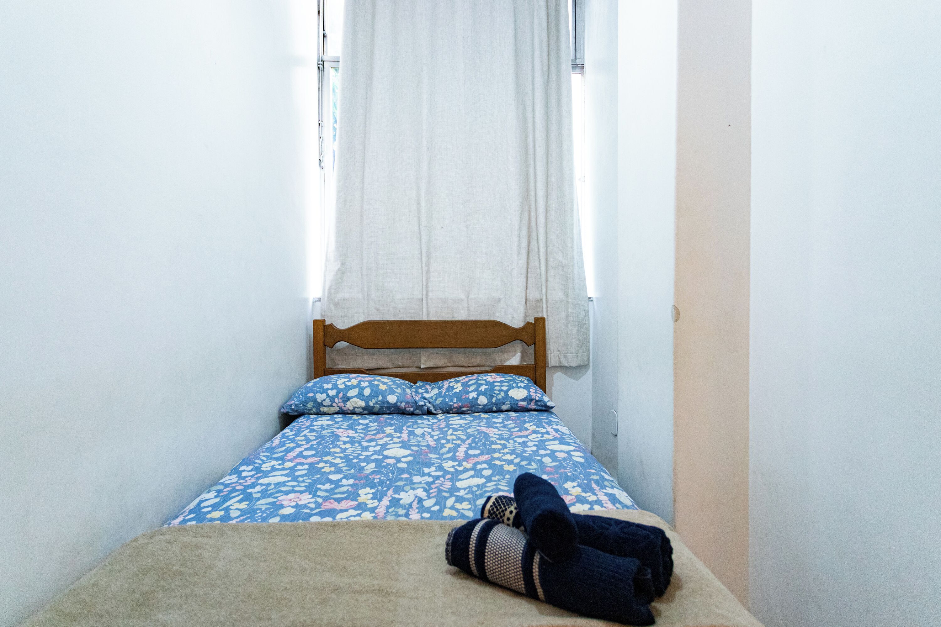A small, narrow room with a single bed, a window with white curtains, and a blanket on the floor.