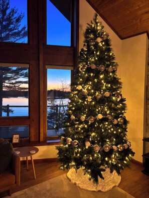 Un sapin de Noël décoré de guirlandes lumineuses et d’ornements, plac é dans une pièce dotée de grandes fenêtres, d’une cheminée et d’un coin salon confortable.