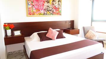 Quarto de hotel com uma cama grande, mesa de cabeceira com um vaso de flores, uma cadeira e uma obra de arte colorida na parede.