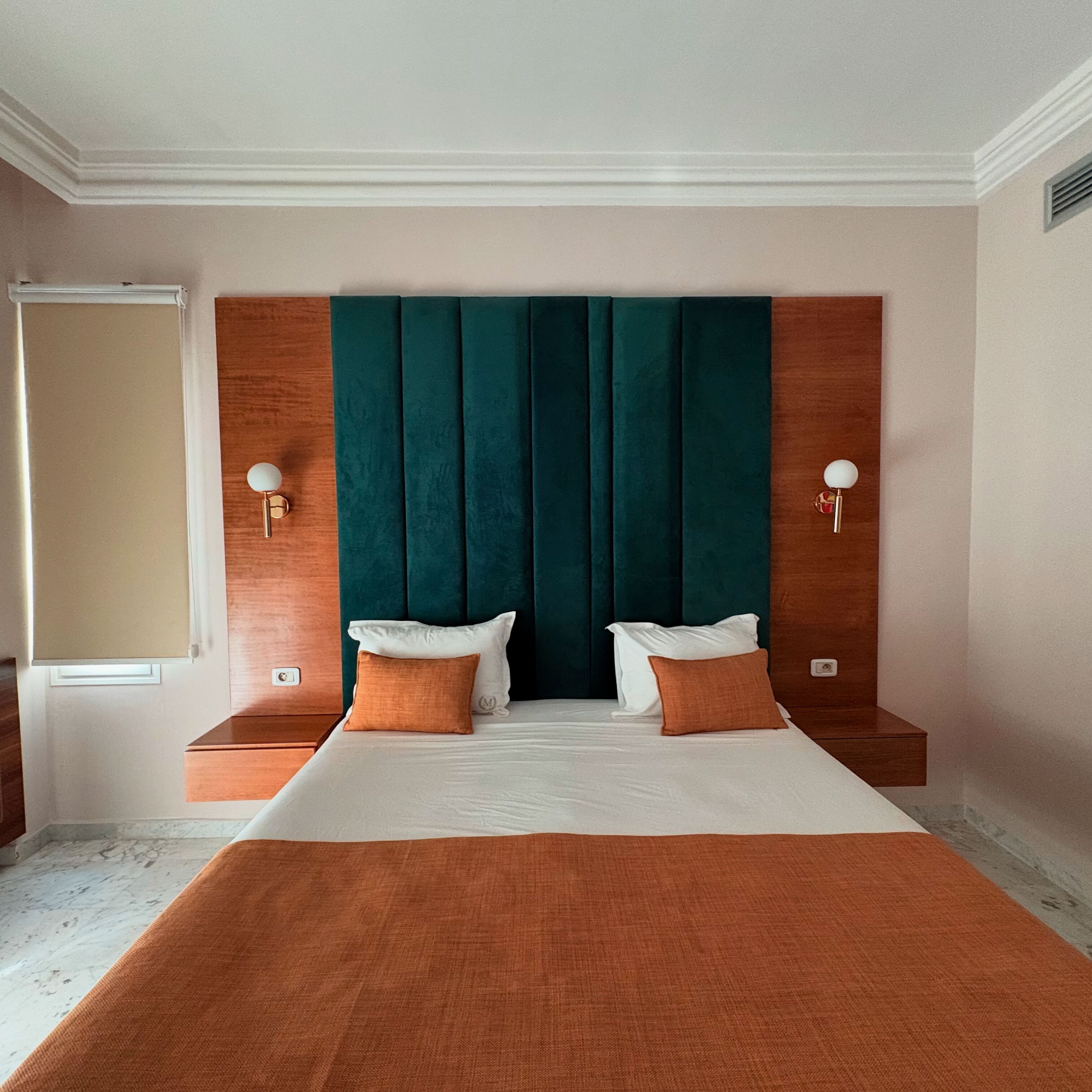 A hotel room with a large bed, two bedside tables, a wooden headboard, and a window with a blind.