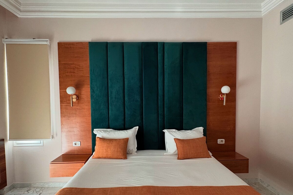 A hotel room with a large bed, two bedside tables, a wooden headboard, and a window with a blind.