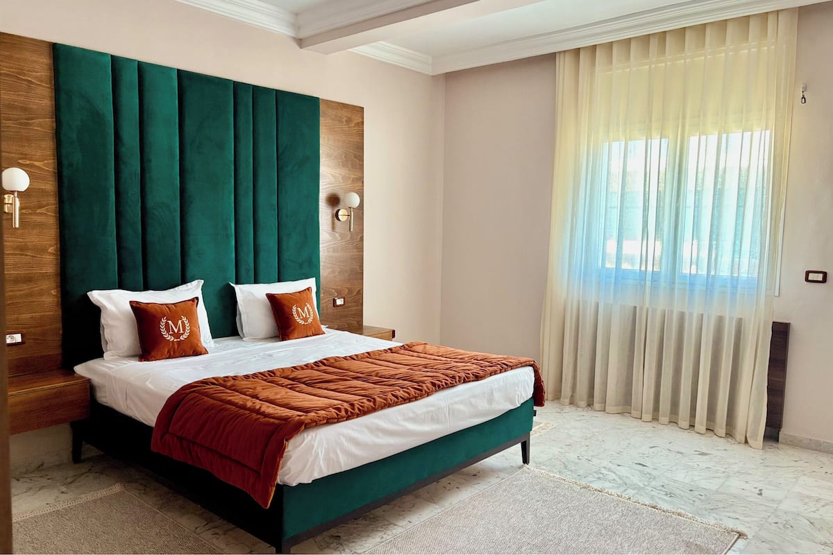 Een netjes opgemaakt slaapkamer met een groot bed, een houten hoofdbord en uitzicht op de omgeving door dunne gordijnen.