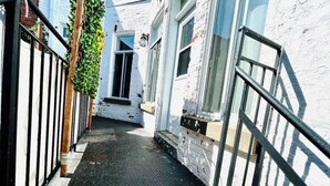 A narrow alleyway with a metal fence, a wooden post, and a building with white walls and a window.