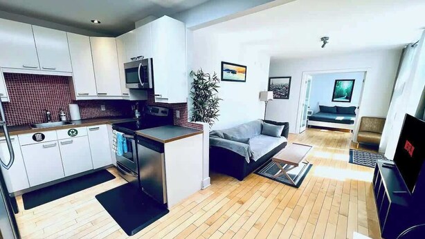 A modern kitchen with white cabinets, a microwave, and a sink. There is a dining area with a sofa and a TV. The room has wooden flooring and a plant.