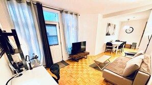 Ein möbliertes Wohnzimmer mit einer Couch, einem Fernseher, einem Schreibtisch mit Kopfhörern und einem Fenster mit Vorhängen.