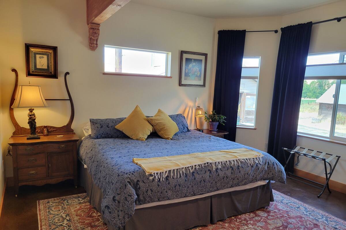 Un dormitorio con una cama grande, una mesita de noche con lámpara, un aparador y una ventana con cortinas.