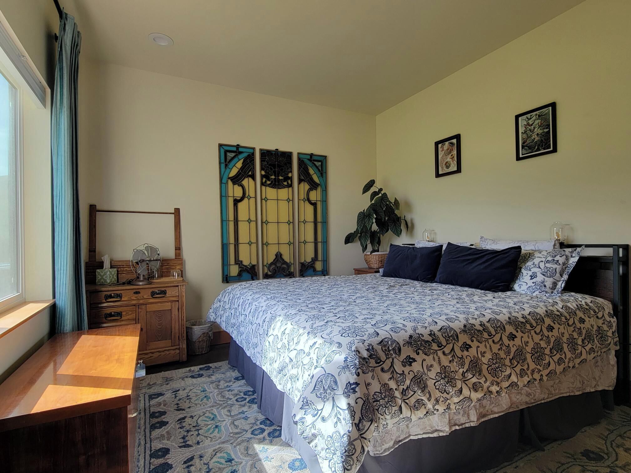 A bedroom with a bed, a desk, a window with curtains, a plant, and two framed pictures on the wall.