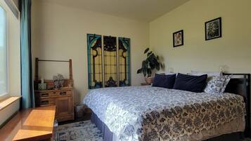A bedroom with a bed, a desk, a window with curtains, a plant, and two framed pictures on the wall.
