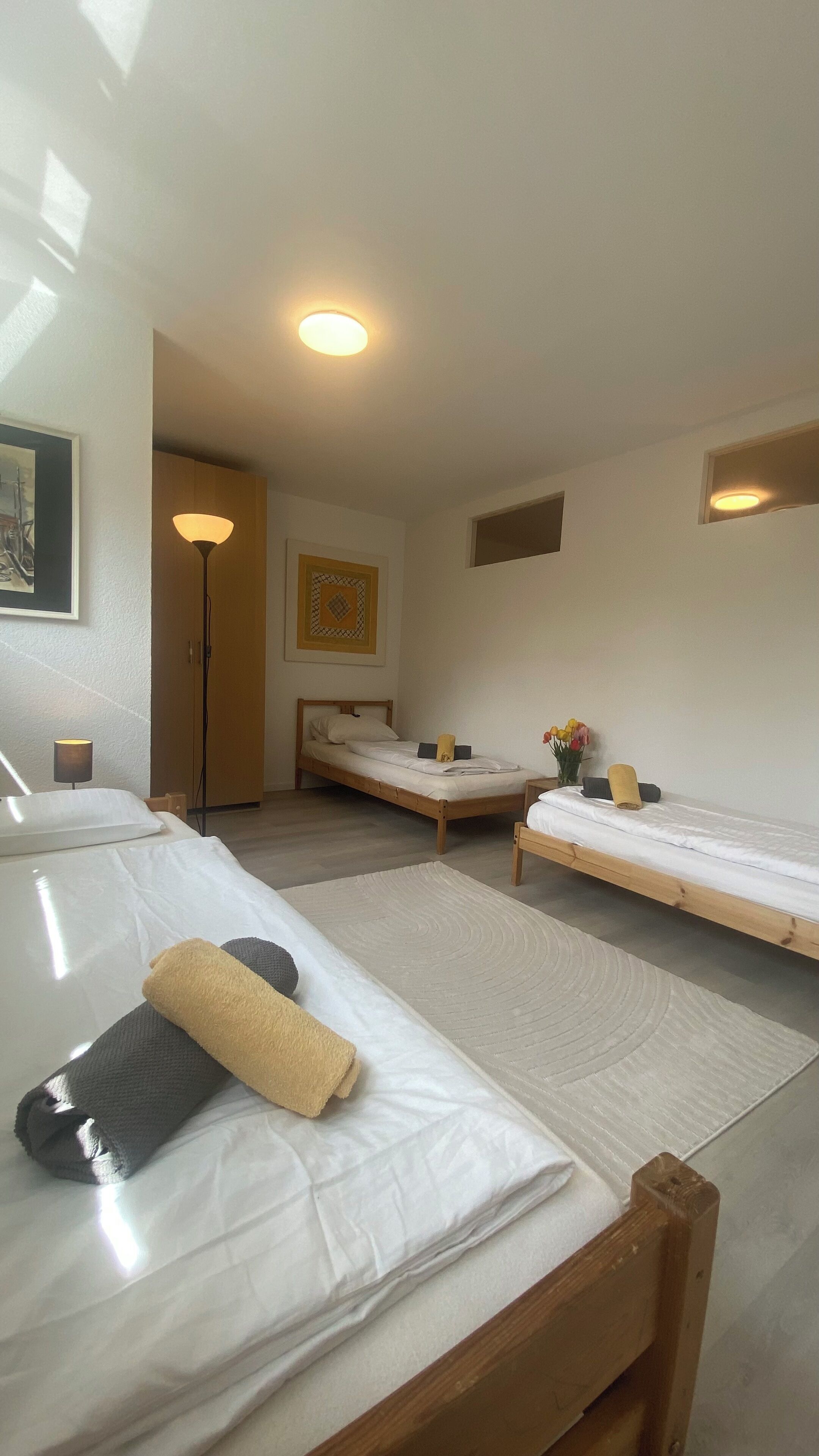 Una camera d'albergo con due letti singoli, un armadio in legno, un comodino con una lampada e una piccola finestra.