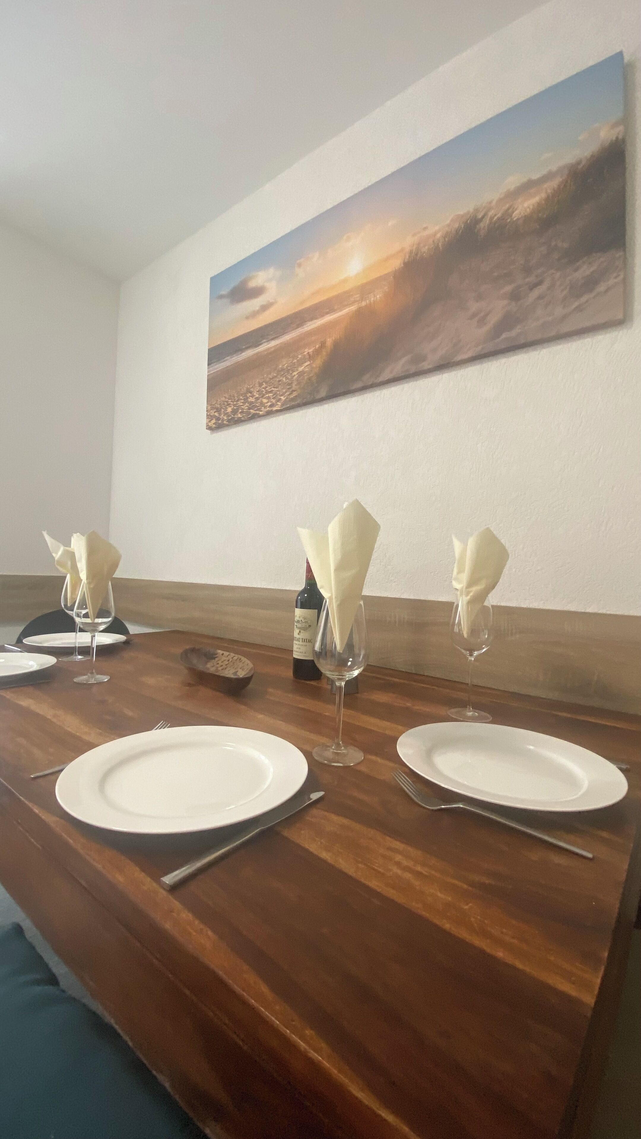 A dining table set with plates, cutlery, and wine glasses, with a painting of a sunset on the wall behind.