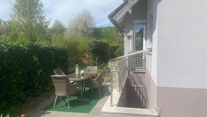En udendørs terrasse med fletmøbler og en grøn måtte, beliggende ved siden af en bygning med hvide vægge og gelænder i metal.