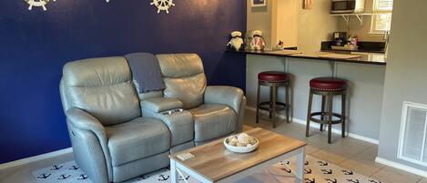 Un salón con un sofá de cuero gris, una mesa de centro de madera, y una zona de cocina con barra alta y taburetes.