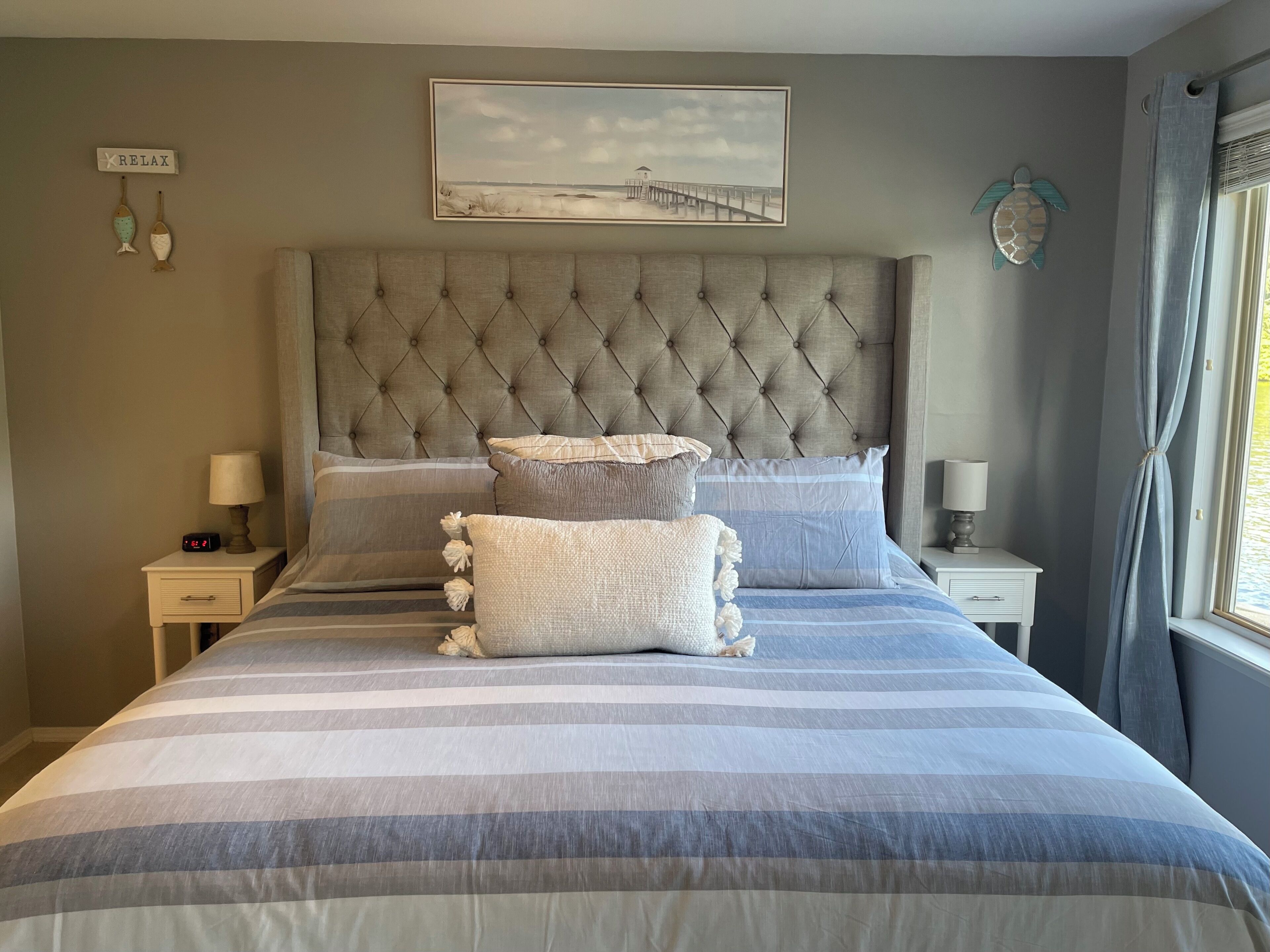 Una camera da letto con un letto grande, biancheria a righe, un'imbottitura a capitonné sulla testiera, un quadro incorniciato sopra il letto e un comodino con una lampada.