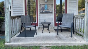 A porch with two chairs and a small table, a red door, and a decorative wall piece.