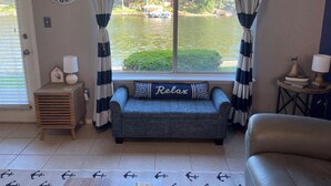 A room with a view of a lake, a sofa with a 'Relax' pillow, a small wooden table with a lamp, and two 'Lifeguard on Duty' signs on the walls.