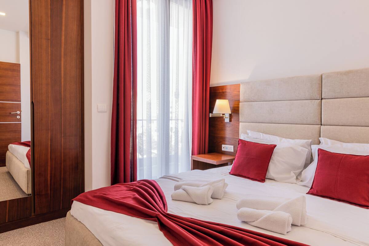 A hotel room with a large bed, red curtains, a bedside lamp, and a wooden wardrobe.