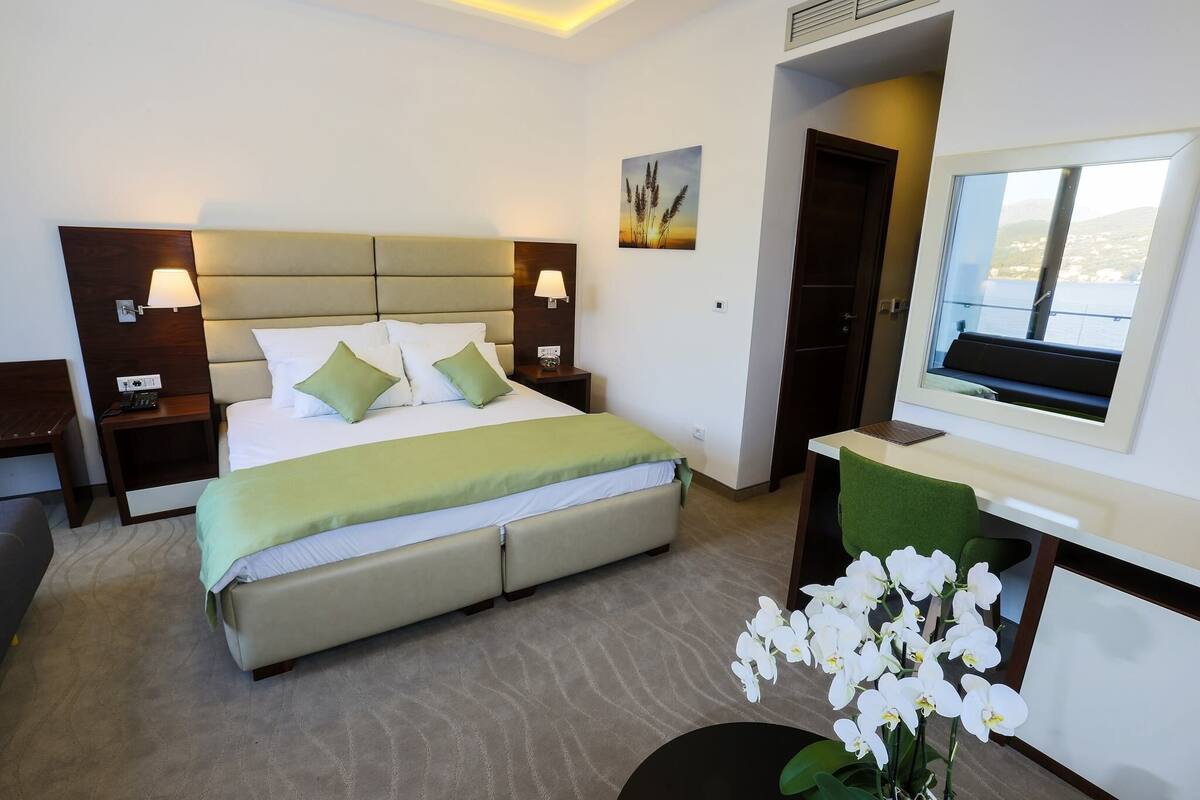 A hotel room with a large bed, two bedside tables with lamps, a desk with a chair, a TV, and a window with a view.