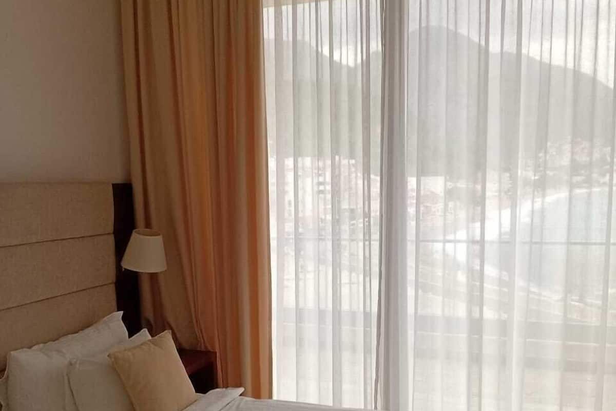 A hotel room with a bed, a nightstand, a lamp, and a view of the outside through sheer curtains.