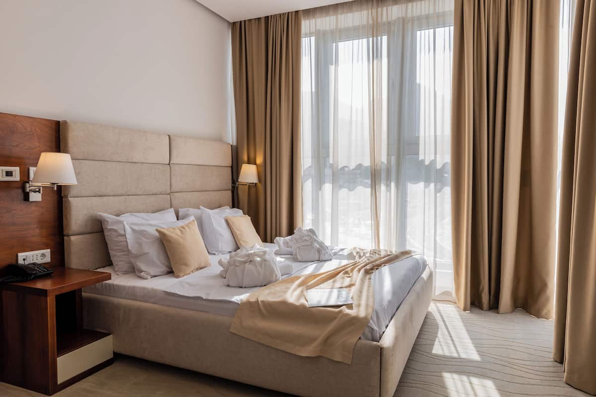 A neatly made bed with white linens, a wooden nightstand with a lamp, and a large window with sheer curtains.