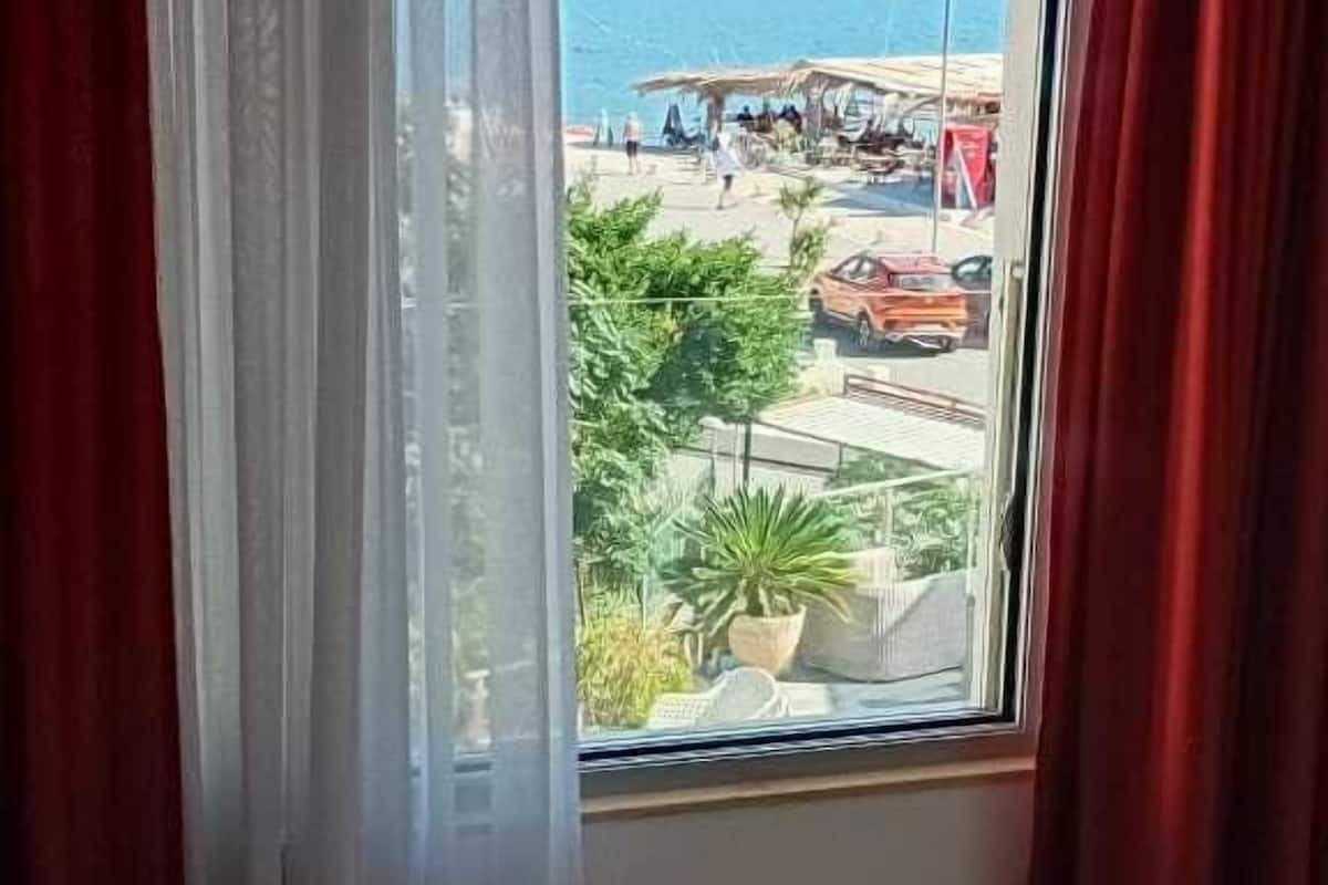 A hotel room with a large window, a bed with white linens, and a view of a beach and mountains.