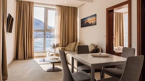 A modern hotel room with a large window, a dining area, and a view of a mountainous landscape. - Porto Sole Hotel (Sutomore)