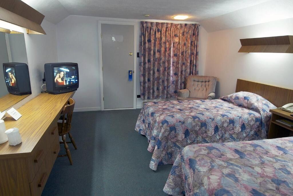 A hotel room with two beds, a desk with a chair, a television, and a chair with a floral bedspread.