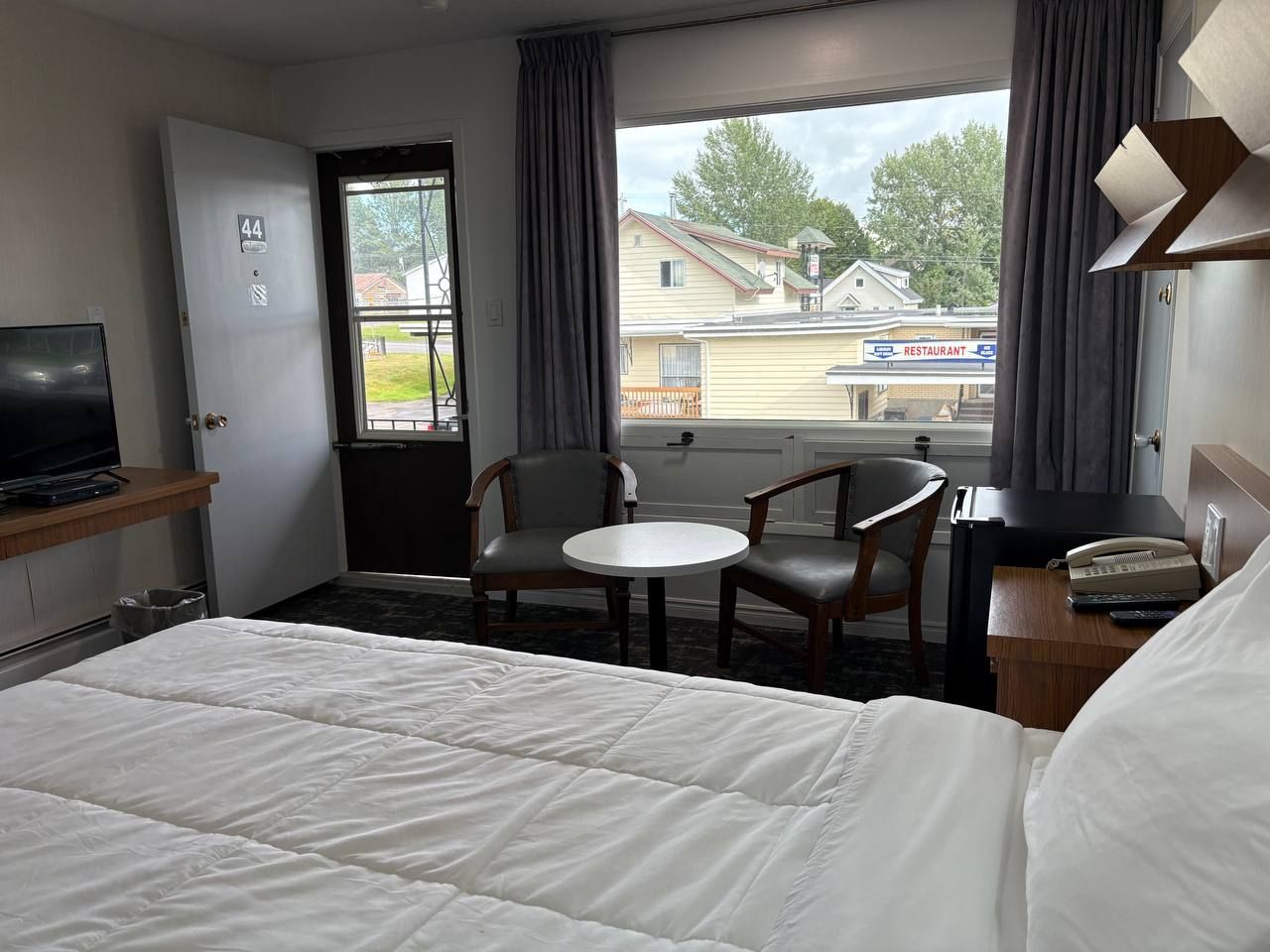 A hotel room with a bed, a desk with a telephone, a chair, a small table, a television on a stand, and a window with curtains.