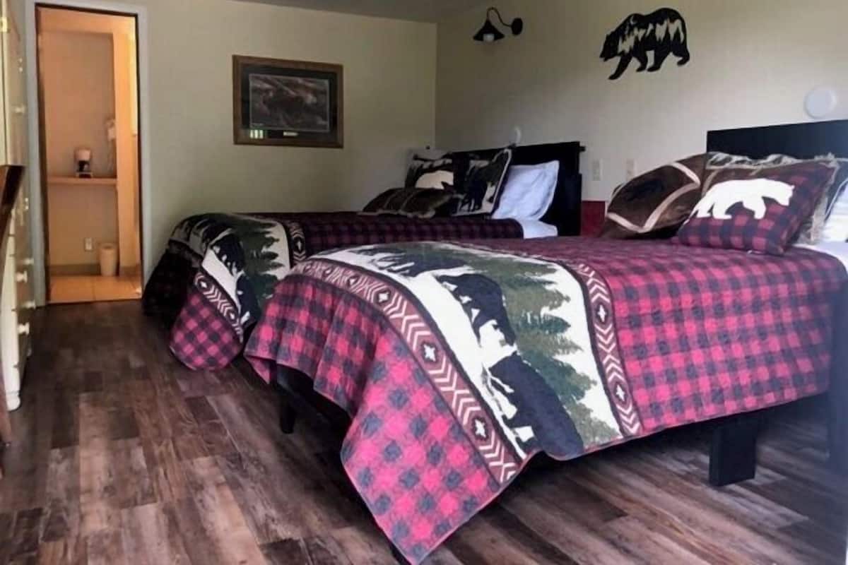 Un dormitorio con dos camas, piso de madera, un cuadro en la pared y una puerta que da a un baño.