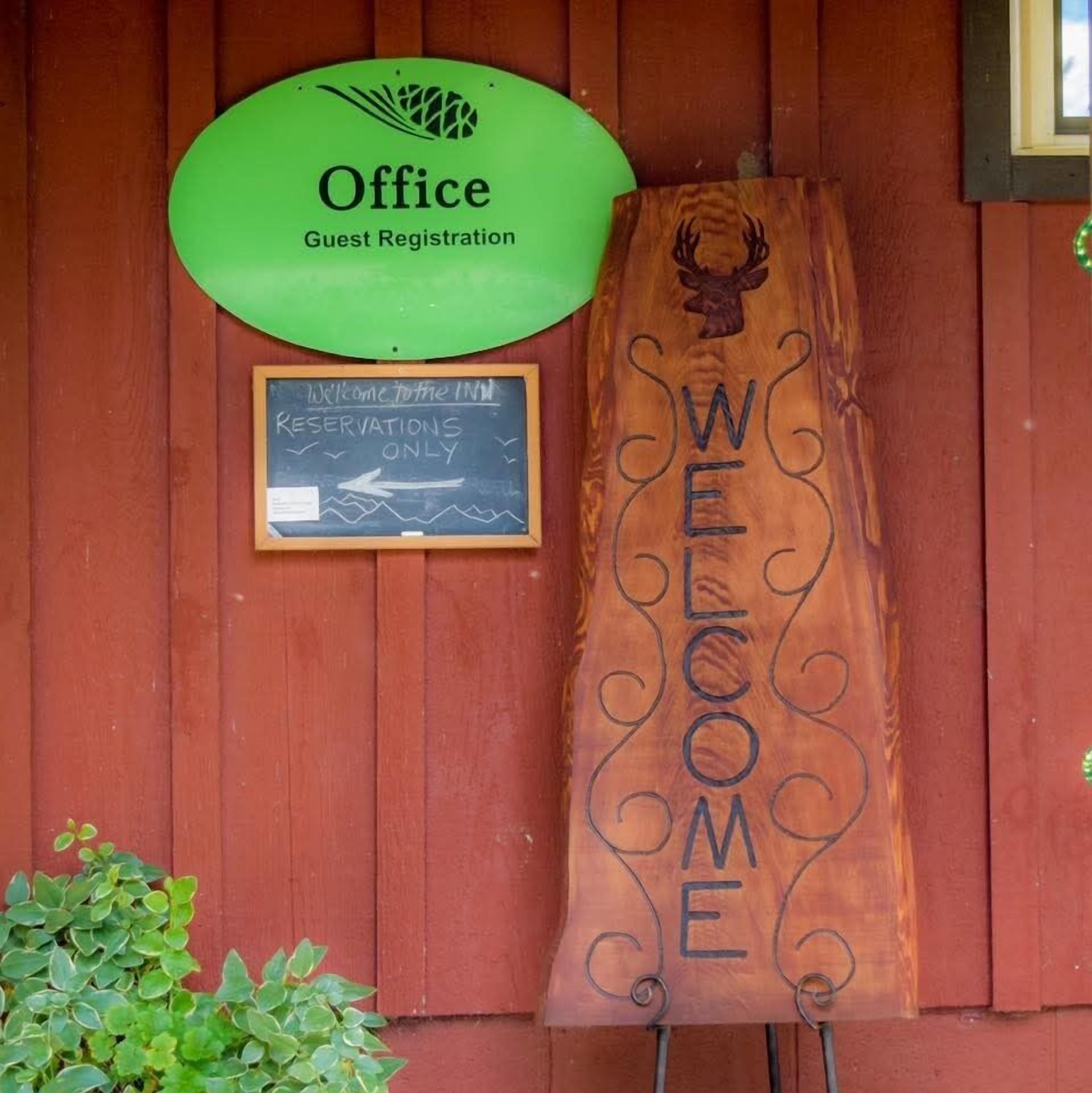 Un letrero de madera con la palabra 'WELCOME' y el logotipo de una cabeza de venado, un letrero verde para registro de huéspedes y una pizarra que indica solo se aceptan reservas.