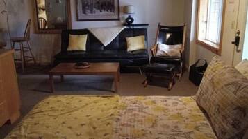 A cozy living room with a sofa, armchair, and a bed. There is a coffee table and a small table with a bowl on it.