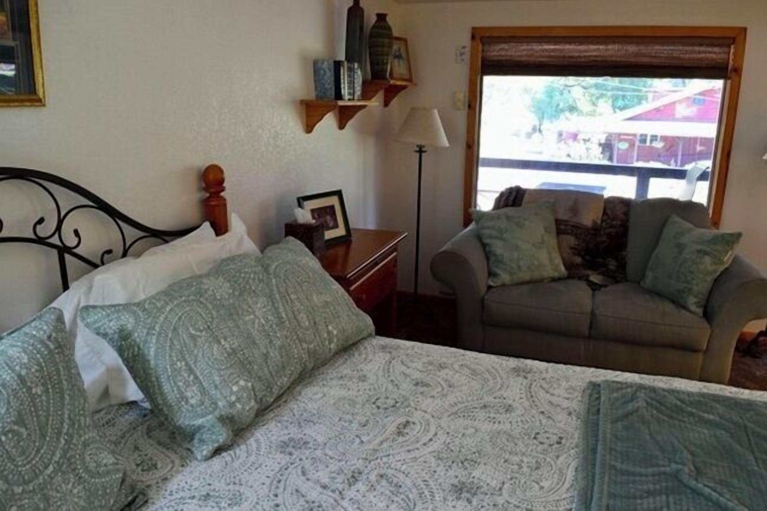 A bedroom with a bed, a sofa, a nightstand, a lamp, and a window with a view of buildings.