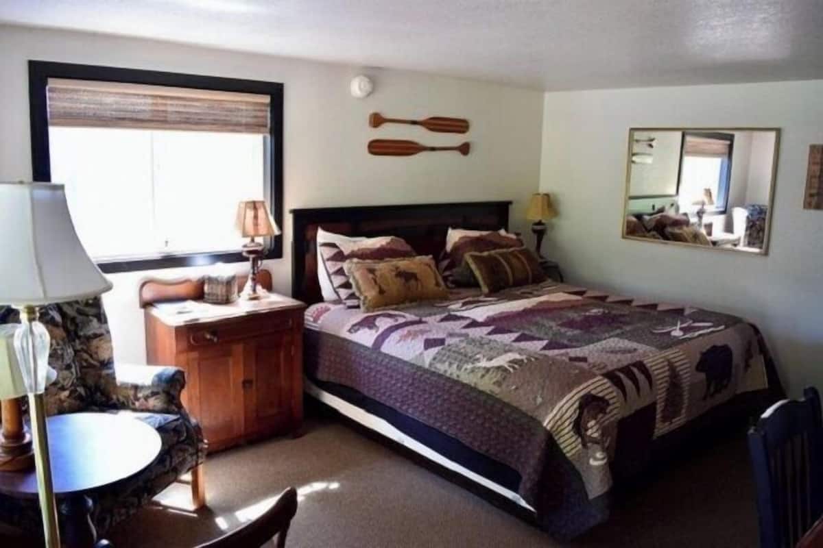Un dormitorio con cama, mesita de noche, lámpara, silla, ventana con persianas y espejo en la pared.
