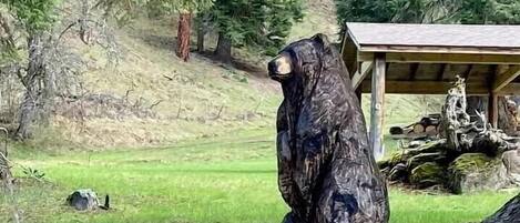 Une sculpture d’ours sur un tronc d’arbre, dans une zone boisée, avec une structure en bois en arrière-plan.