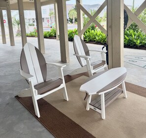Two white Adirondack chairs with wooden slats and a bench, placed on a concrete patio with a brown mat.