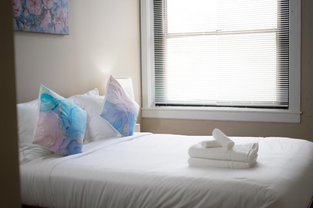 A neatly made bed with white linens and a folded towel, a window with blinds, and a floral painting on the wall.
