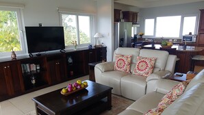 A spacious living room with a large sofa, a coffee table with fruit, a flat-screen TV, and a kitchen in the background. - Playa Hermosa Ocean View Estate (Playa Hermosa)