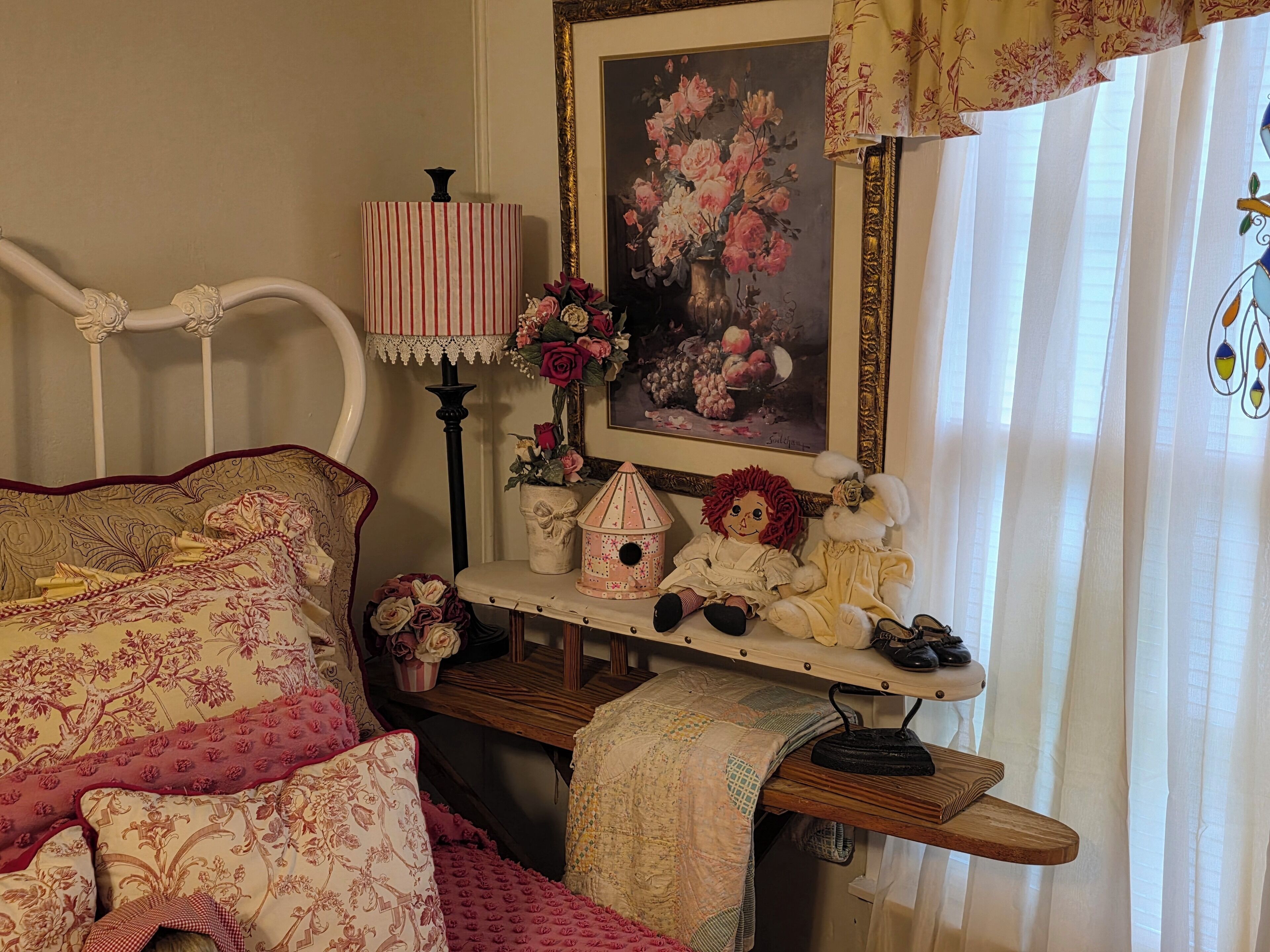 A cozy bedroom with a bed, a bedside table with a lamp, a framed floral painting, and a small shelf with decorative items.