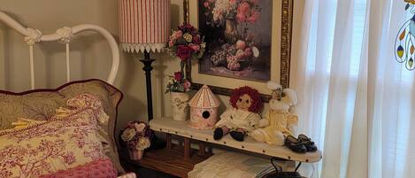 A cozy bedroom with a bed, a bedside table with a lamp, a framed floral painting, and a small shelf with decorative items.