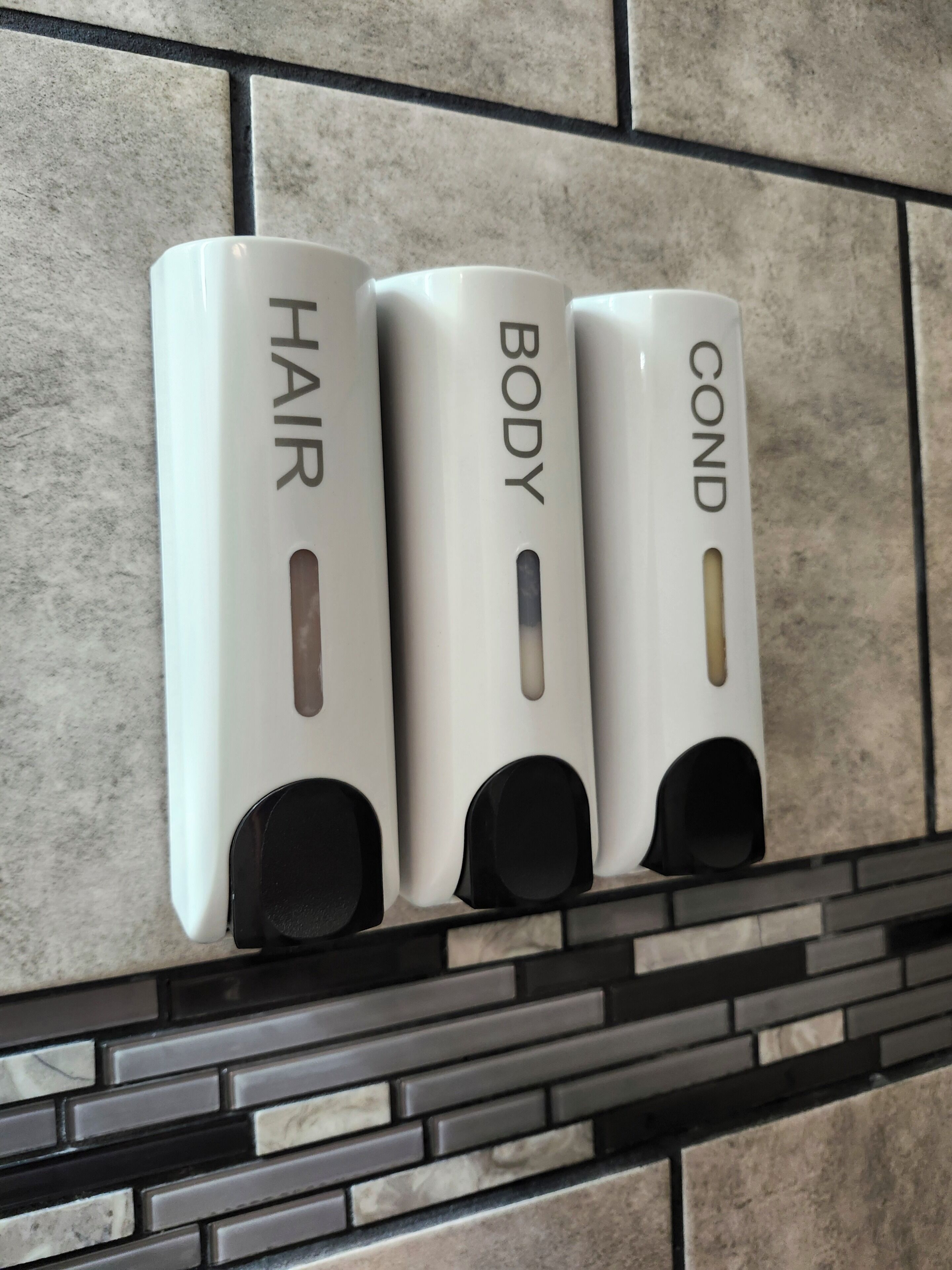 Three white dispensers labeled 'HAIR,' 'BODY,' and 'COND' on a tiled wall.