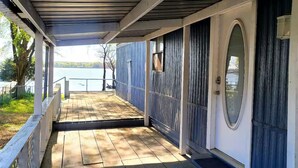 Varanda coberta com deck de madeira, porta branca com painel de vidro oval e vista para um lago.