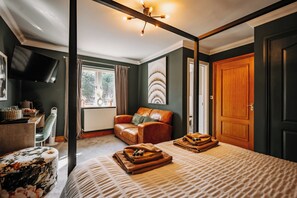 A bedroom with a four-poster bed, a leather armchair, a desk with a flat-screen TV, and a large window with curtains.