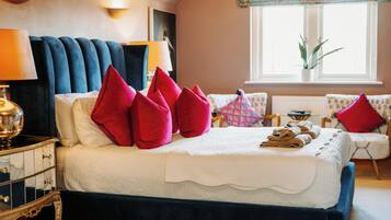 Una cama bien hecha con sábanas blancas y almohadas rojas, una mesita de noche con una lámpara, y vistas a una ventana con una planta.