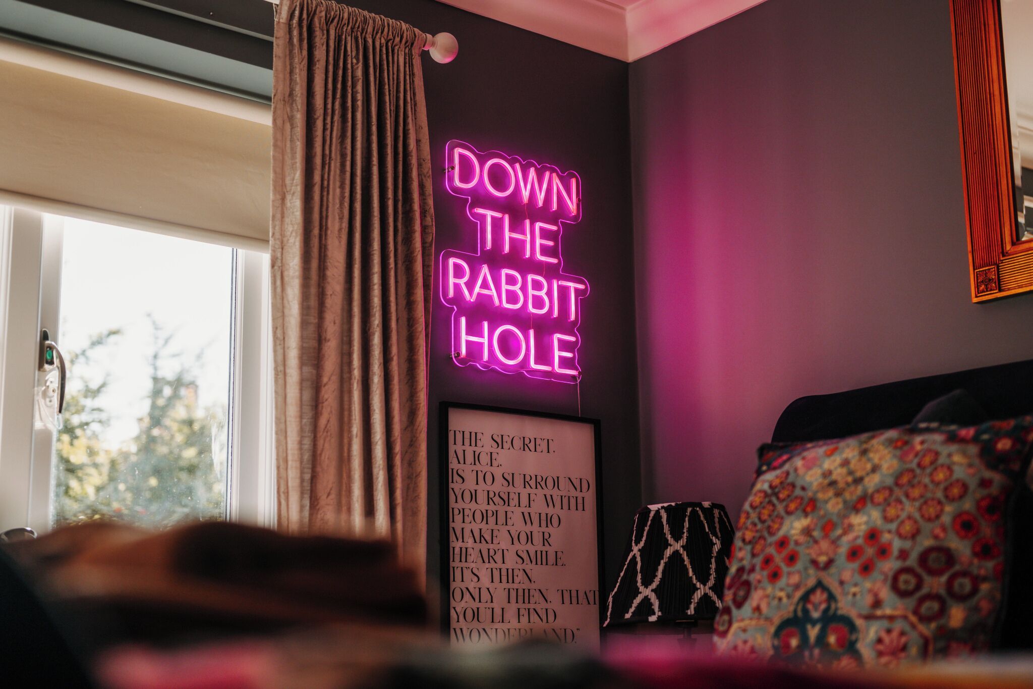 A room with a neon sign reading 'DOWN THE RABBIT HOLE' and a framed quote on the wall.