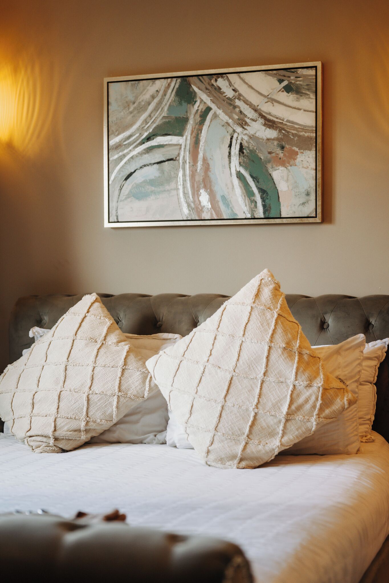A bedroom with a tufted headboard, two large pillows, and a framed abstract painting on the wall.