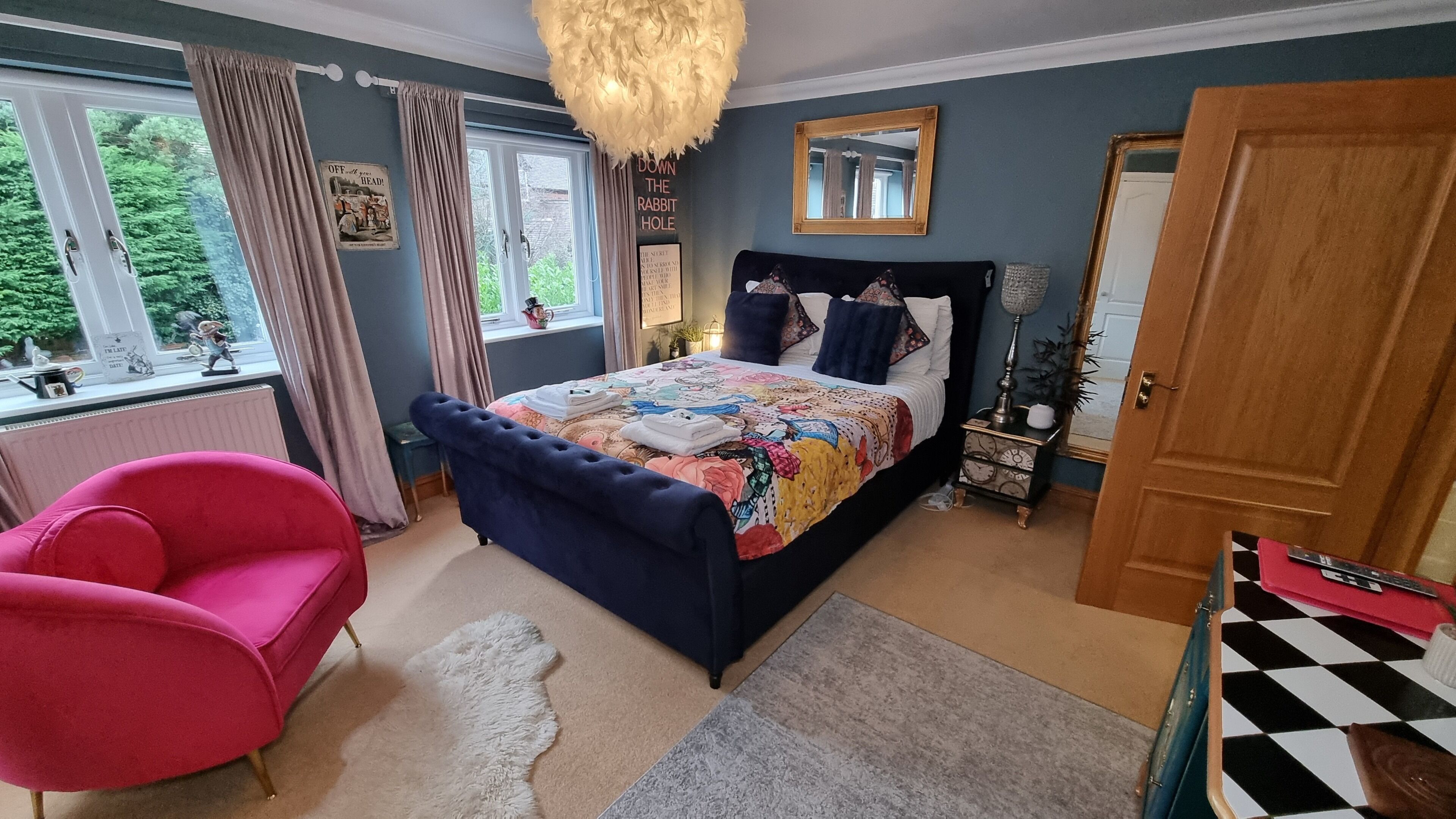 Un dormitorio con una cama grande, una silla rosa, un aparador y un ventanal con cortinas.