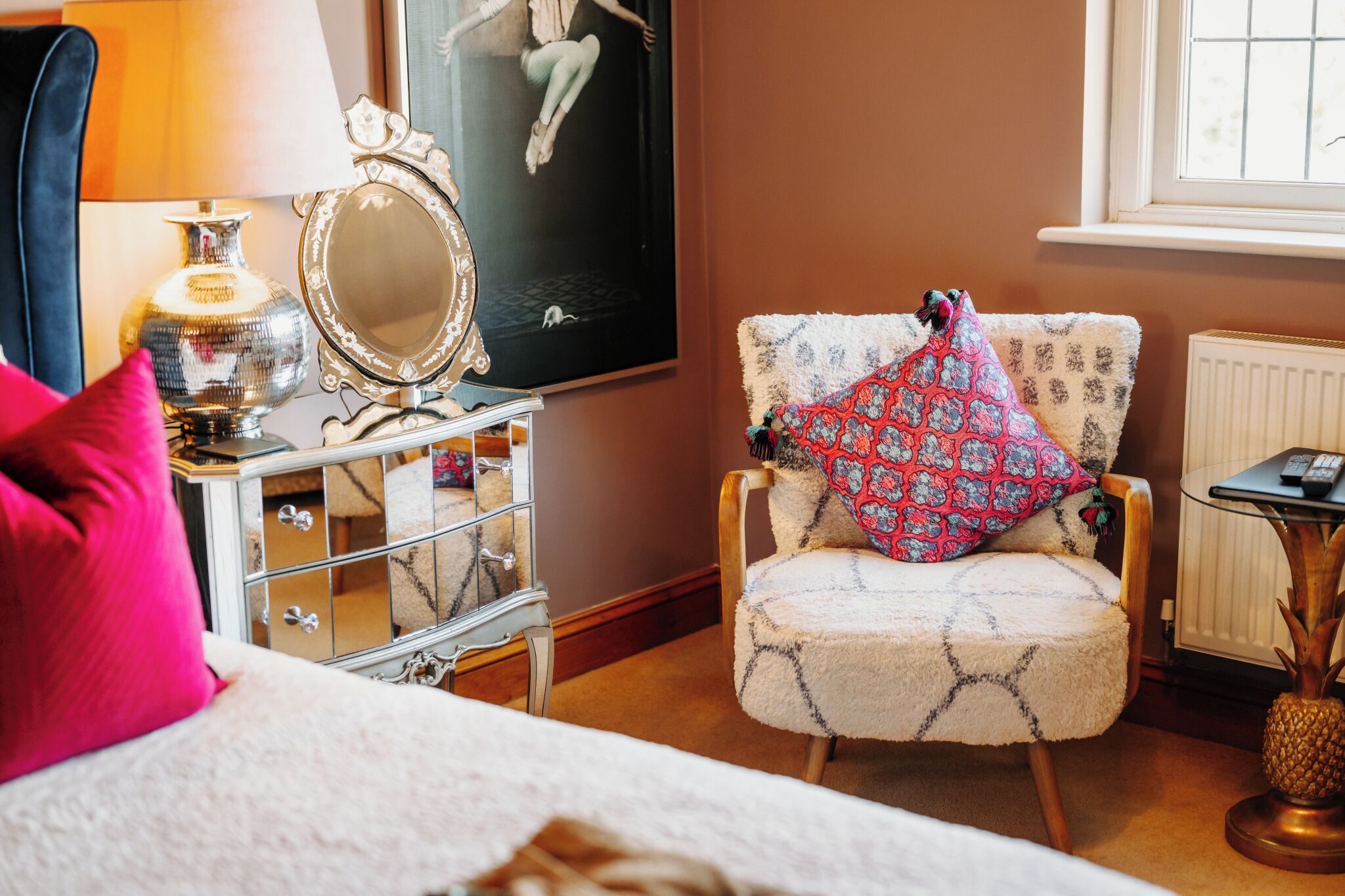 A bedroom with a bed, a dresser with a mirror, a chair with a patterned pillow, a lamp, and a poster on the wall.