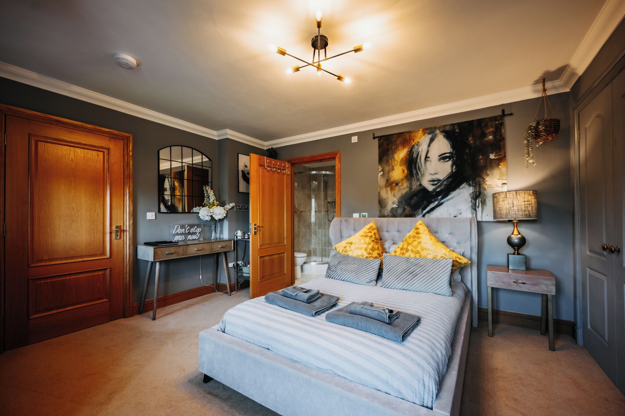 A bedroom with a large bed, a vanity with a mirror, a painting on the wall, and a chandelier.
